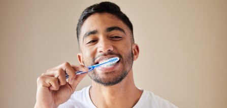 Man in white t-shirt brushing teeth in front of tan background