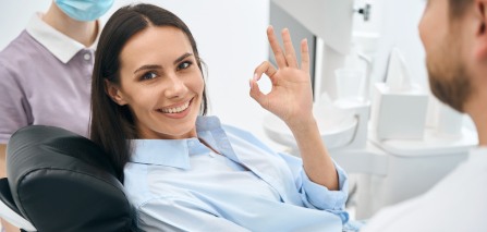 Woman in blue shirt in dental chair making “ok” sign with one hand